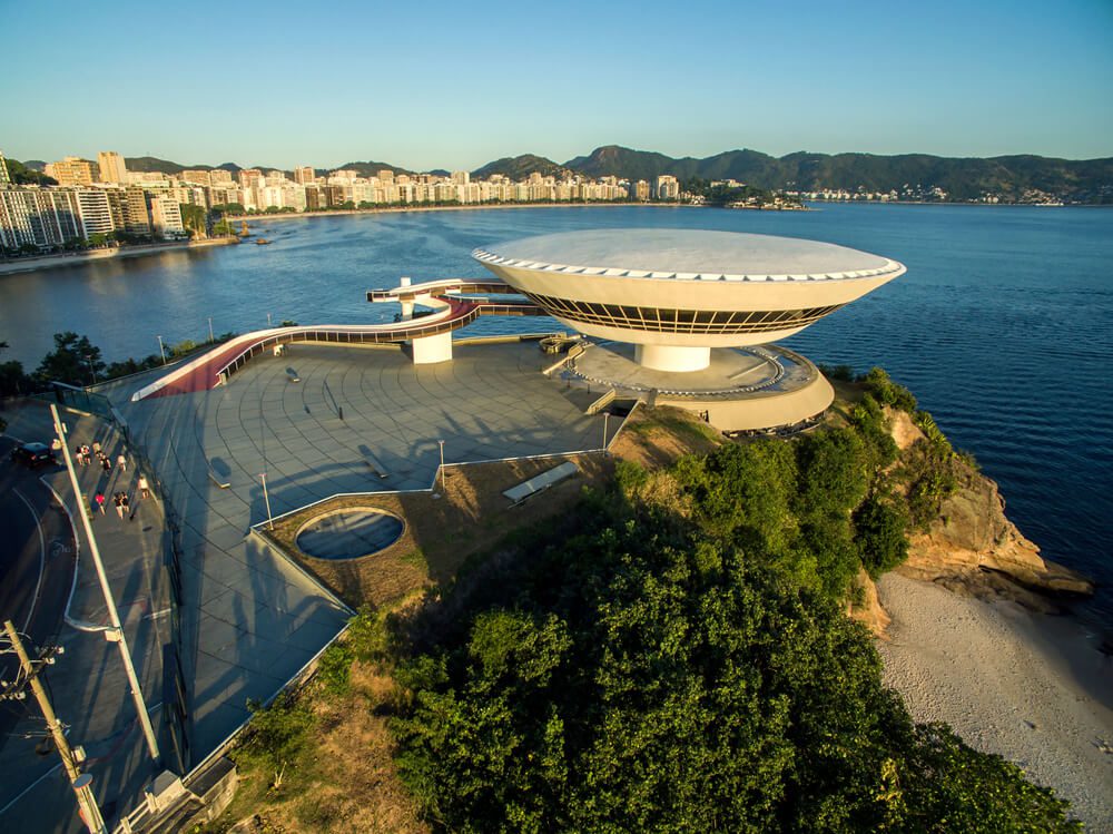 Vista aérea do Museu de Arte Contemporânea de Niterói, um edifício futurista em forma de disco com vista para a água em uma das melhores cidades do Rio de Janeiro, com paisagem urbana e montanhas ao fundo.
