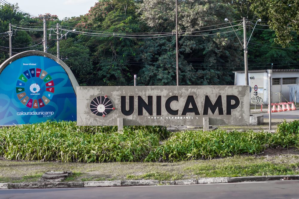 Uma placa de concreto com o texto UNICAMP se destaca entre plantas verdes. Ao fundo, um painel circular colorido e árvores marcam a entrada do campus universitário — um cenário convidativo para quem deseja morar em Campinas.