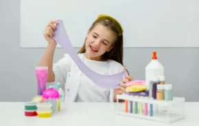 Imagem de uma menina sorridente, vestindo um jaleco branco e óculos de proteção amarelos, estica um slime roxo em uma mesa com frascos coloridos, tintas e recipientes, para ilustrar matéria sobre como fazer slime caseiro.