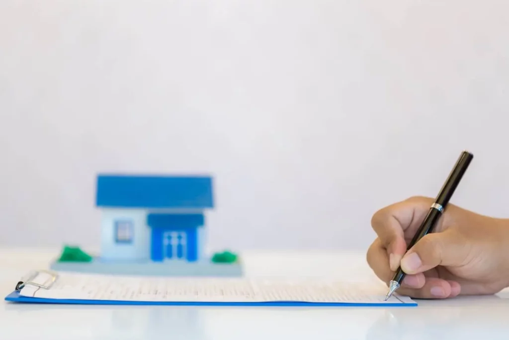 Imagem de uma pessoa assinando um documento em uma prancheta que está em cima de uma mesa ao lado de uma casa de madeira em tamanho miniatura para ilustrar matéria sobre o que é certidão de ônus reais de imóveis
