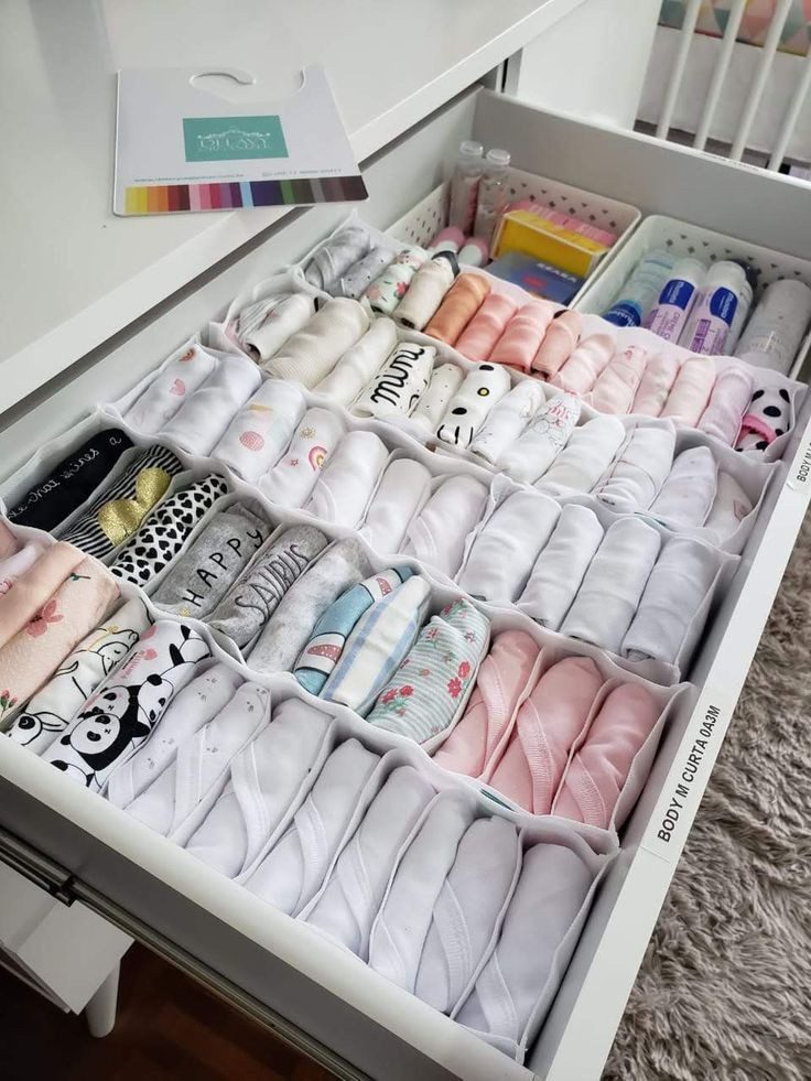 Uma gaveta bem organizada mostra como montar um enxoval de bebê, com divisórias para guardar roupinhas de bebê enroladas, meias e mamadeiras. Tudo é separado por cor e tipo para um arranjo organizado e visualmente agradável.