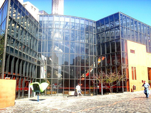 Um edifício moderno com grandes janelas de vidro reflete o céu e a paisagem ao redor. A fachada apresenta linhas angulares, sugerindo o design inovador frequentemente visto em museus de Curitiba. Algumas pessoas caminham pelo pátio de paralelepípedos em frente.