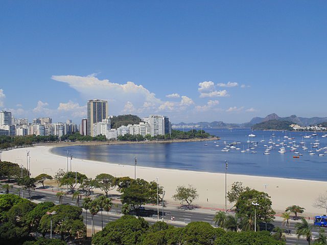 Uma praia de areia curva, ladeada por árvores e uma estrada, com prédios altos e morros ao fundo, perto da estação de metrô Jardim Oceânico, com vários barcos ancorados nas águas azuis e calmas sob um céu limpo.