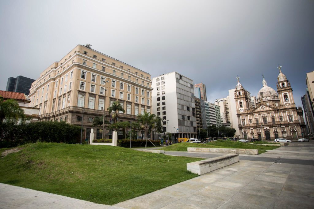 Uma praça com grama verde, palmeiras, edifícios modernos e uma grande igreja histórica com duas cúpulas e arquitetura ornamentada sob um céu nublado — um cenário convidativo para aqueles que sonham com Morar no Rio de Janeiro.