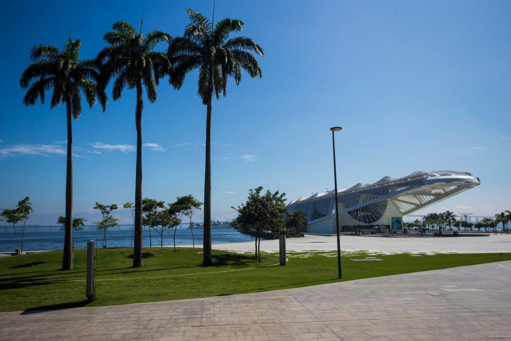Edifício branco moderno com design futurista próximo à orla, cercado por palmeiras e áreas gramadas sob um céu azul claro — perfeito para quem sonha em morar no Rio de Janeiro.