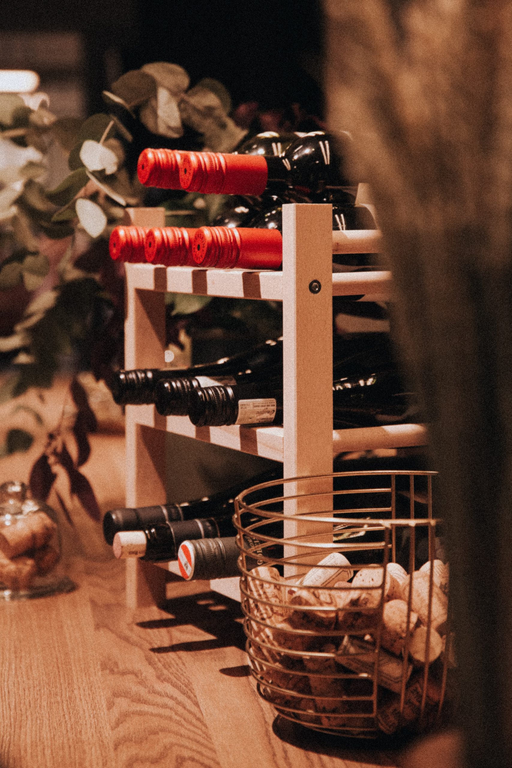 Uma adega de madeira abriga várias garrafas de vinho com tampas vermelhas e pretas — um cenário convidativo para quem está aprendendo a montar uma adega. À frente, uma cesta de metal transborda de rolhas, iluminada de forma acolhedora entre folhas e decoração ao fundo.