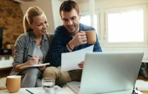 Imagem de um casal sorridente sentado juntos em casa, olhando para alguns documentos e um notebook para ilustrar matéria que responde se é melhor alugar ou vender imóvel. A mulher segura um caderno e uma caneta, enquanto o homem segura uma caneca. A luz do sol entra pelas janelas ao fundo.