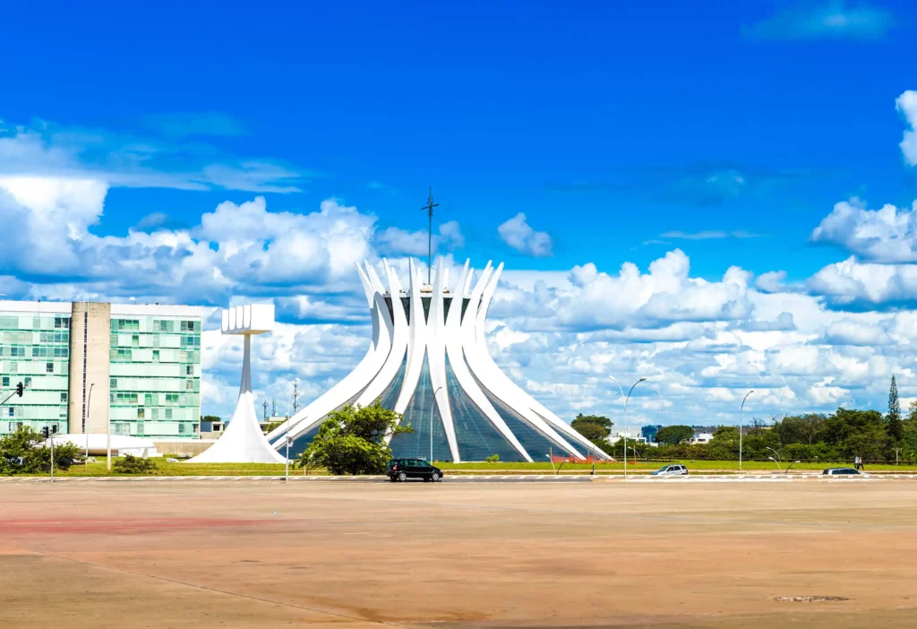 Imagem da moderna Catedral de Brasília, branca e em forma de coroa, contra um céu azul brilhante com nuvens espalhadas - uma atração imperdível quando se explora o que fazer em Brasília, cercada por edifícios modernos e uma área pavimentada quase vazia