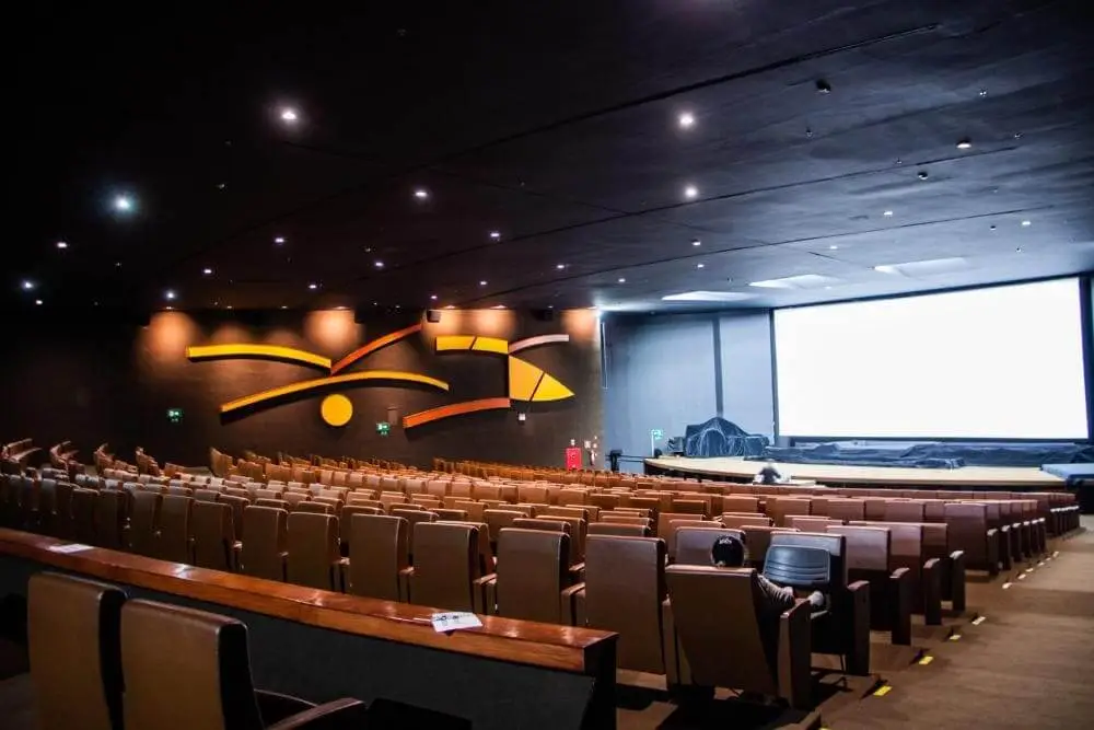 Imagem de uma sala de cinema do Cine Brasília, em Brasília, mostra um grande e moderno auditório de cinema com poltronas marrons vazias, uma tela bem iluminada, paredes escuras e obras de arte abstratas em laranja na parede lateral