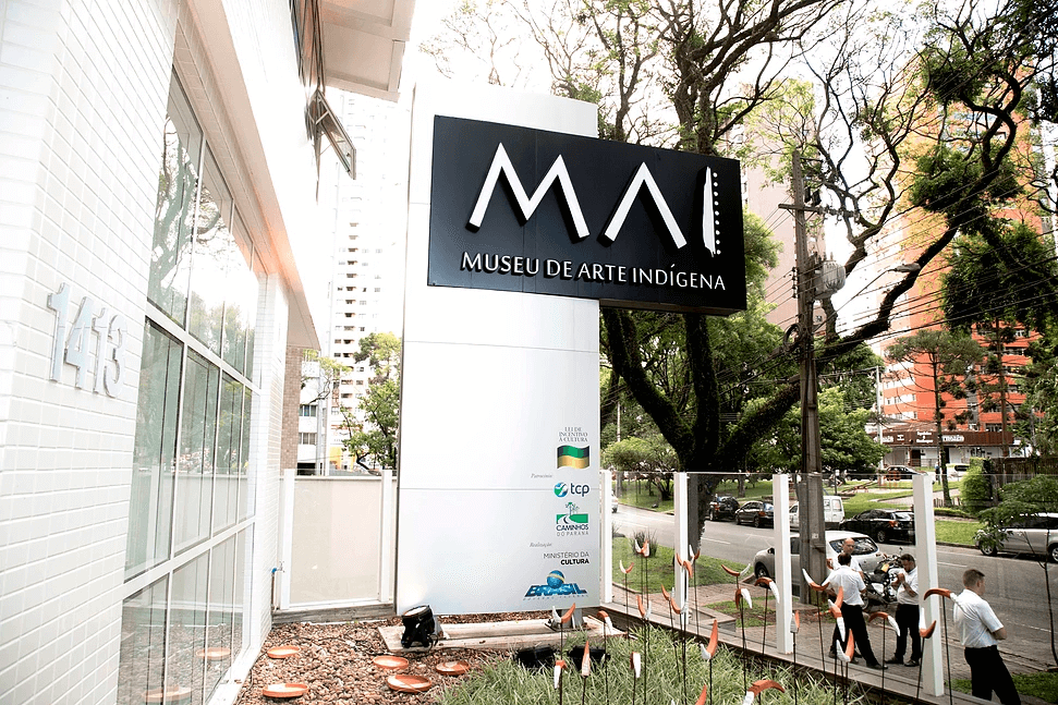 Uma grande placa com os dizeres “MAI Museu de Arte Indígena” fica do lado de fora de um moderno edifício branco com janelas de vidro; entre os museus de Curitiba, ele está situado em uma rua movimentada, ladeada por árvores, prédios altos e transeuntes.