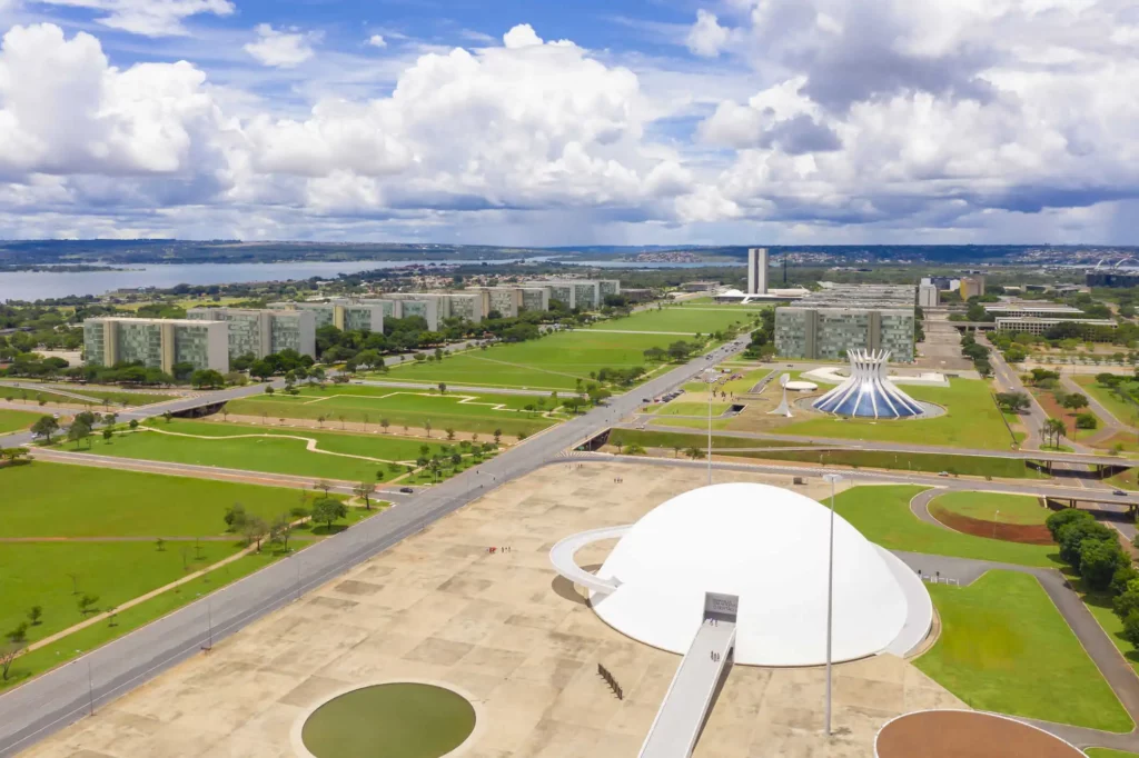 Imagem da vista aérea de Brasília, Brasil, destacando os prédios governamentais modernistas, a cúpula branca do Museu Nacional, a Catedral de Brasília e os gramados verdes
