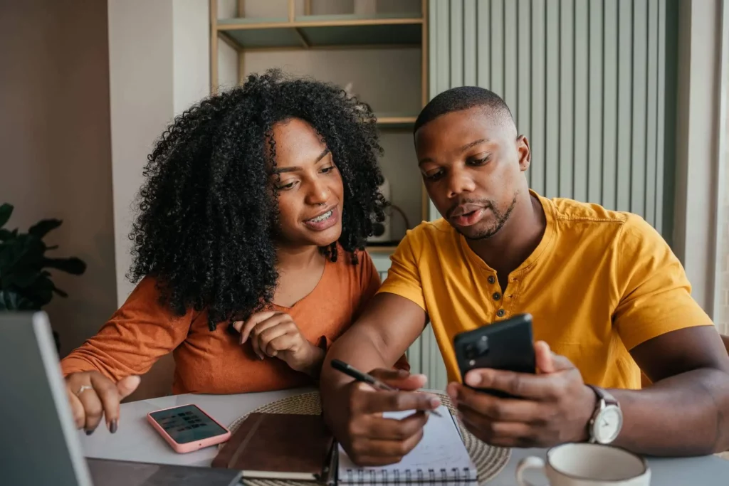 Imagem de um casal formado por um homem e uma mulher sentados na mesa de casa analisando informações em um computador e um celular e fazendo anotações em um papel para ilustrar matéria sobre o que é imposto de renda sobre aluguel
