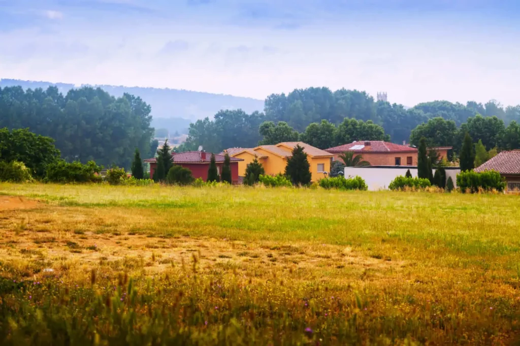 Imagem de um campo gramado se estende em direção a várias casas com telhados vermelhos e amarelos, cercadas por árvores verdes e colinas distantes sob um céu azul e ligeiramente nublado, para ilustrar matéria sobre o que é o NIRF do imóvel rural