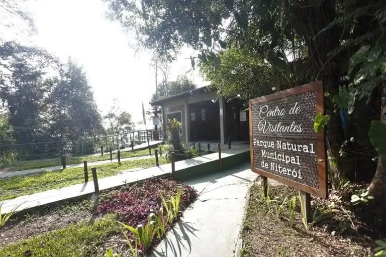Imagem da entrada do Parque Natural Municipal de Niterói mostra um pequeno prédio cercado por árvores e plantas com uma placa de madeira na frente onde se lê Centro de Visitantes Parque Natural Municipal de Niterói