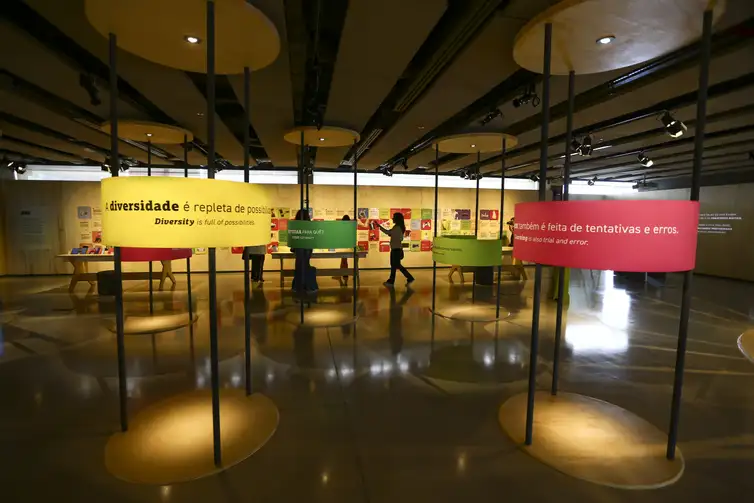 Imagem de um espaço de exposições dentro do Sesi Lab Brasília mostra banners coloridos e bilíngues exibindo textos em português e inglês