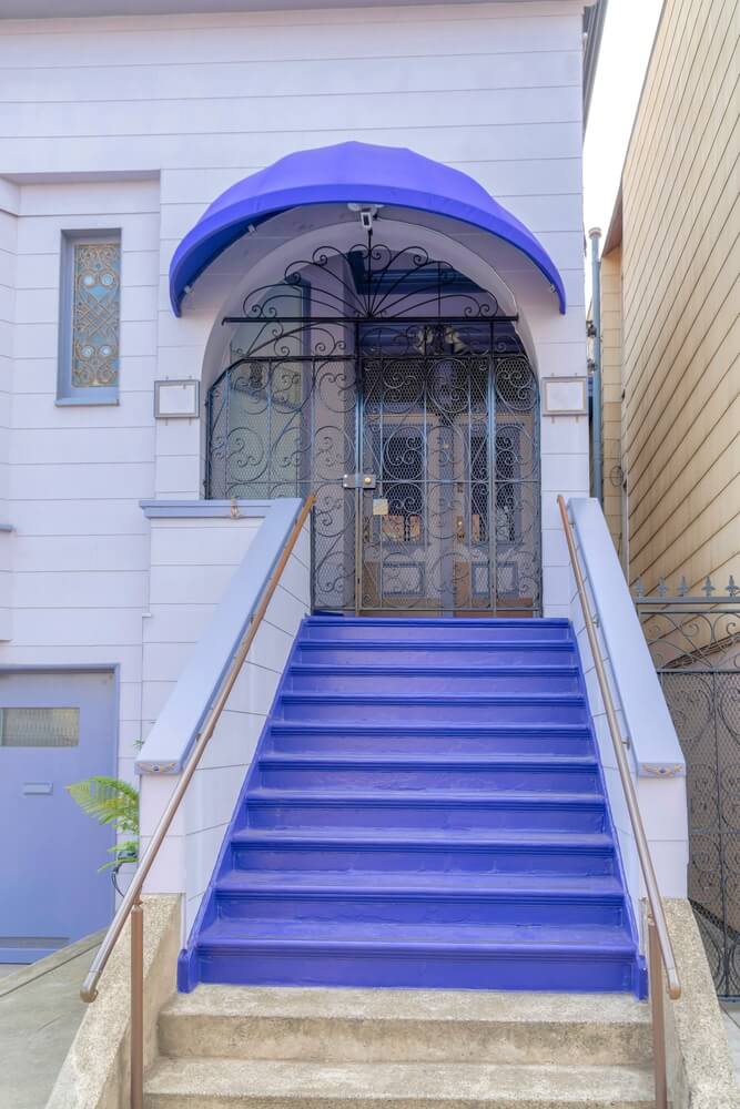 Uma escada pintada em roxo vibrante leva até a entrada de um edifício com portas de metal ornamentadas e um toldo roxo combinando, criando uma charmosa fachada em contraste com o exterior de cor clara com janelas decorativas.