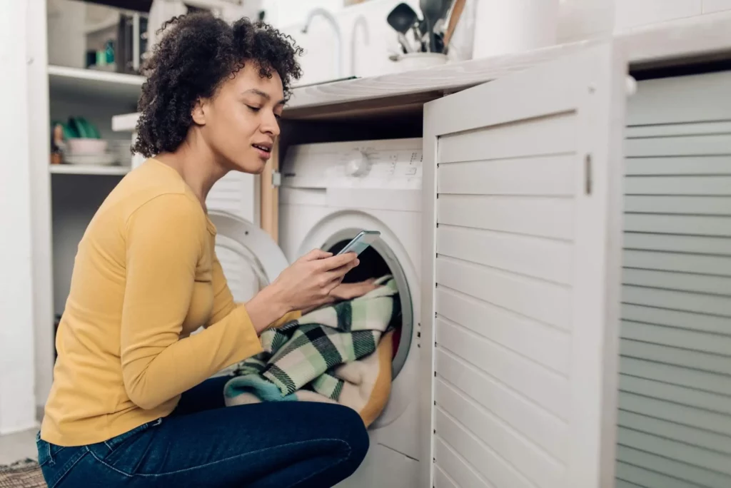 Imagem de uma mulher colocando roupas para lavar enquanto lê alguma informação no celular para ilustrar matéria sobre como limpar máquina de lavar com vinagre e bicarbonato