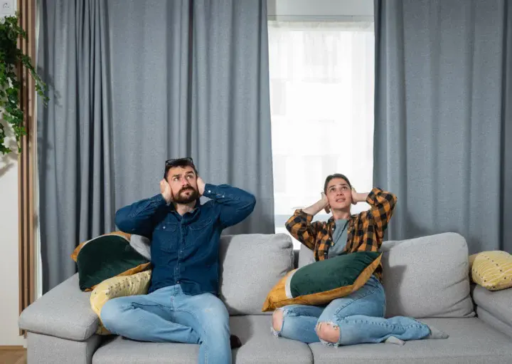 Imagem de um casal sentado no sofá de casa com as mãos na orelha olhando para cima para ilustrar matéria sobre a lei da perturbação do sossego