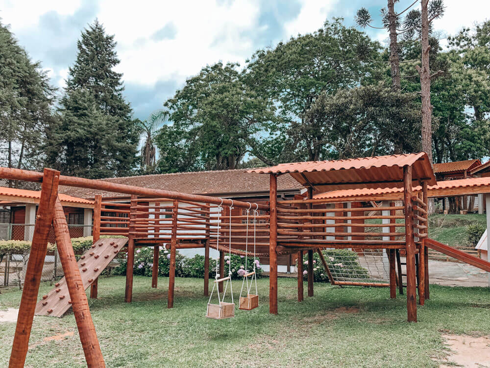 Uma estrutura de playground de madeira com balanços, parede de escalada e escada fica na grama em frente a prédios e árvores altas sob um céu nublado, típico de escolas particulares em Cotia.