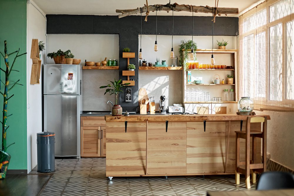 A imagem mostra uma cozinha americana rústica com conceito bem aberto. Nela há uma bancada e diferentes estantes de madeira. Há também um lustre feito de um tronco de árvore, além de geladeira de inox, pia e diferentes utensílios (temperos, cafeteira, copos, etc).