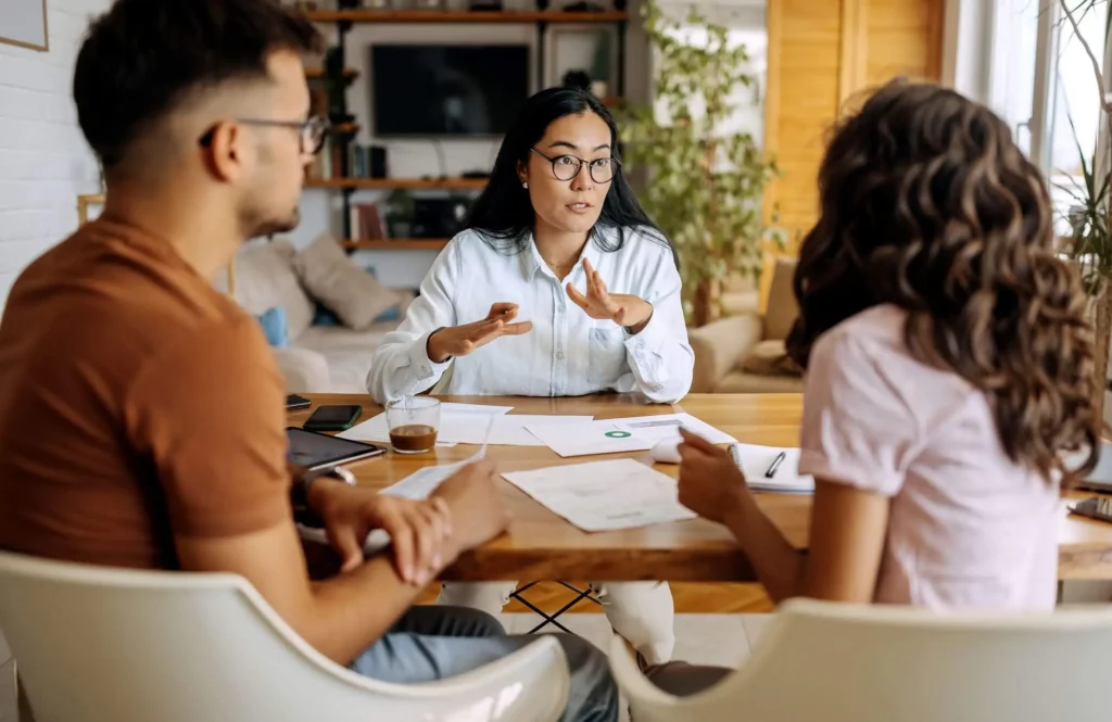 Imagem de uma mulher sentada em uma mesa discutindo a papelada da hipoteca de imóvel com um homem e uma mulher, gesticulando com as mãos. Documentos, um telefone e uma xícara de café estão sobre a mesa em uma sala aconchegante e bem iluminada com plantas e prateleiras