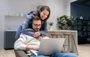 Imagem de uma mulher sorridente abraçando um homem por trás enquanto ele está sentado em uma cadeira, usando um notebook e fones de ouvido em uma cozinha moderna, para ilustrar matéria sobre como escolher inquilino