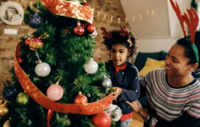 Imagem de uma mulher e uma criança usando bandanas festivas decorando uma árvore de Natal com enfeites e fitas vermelhas em uma sala aconchegante e bem iluminada, compartilhando dicas sobre como montar uma árvore de Natal enquanto a criança se concentra atentamente