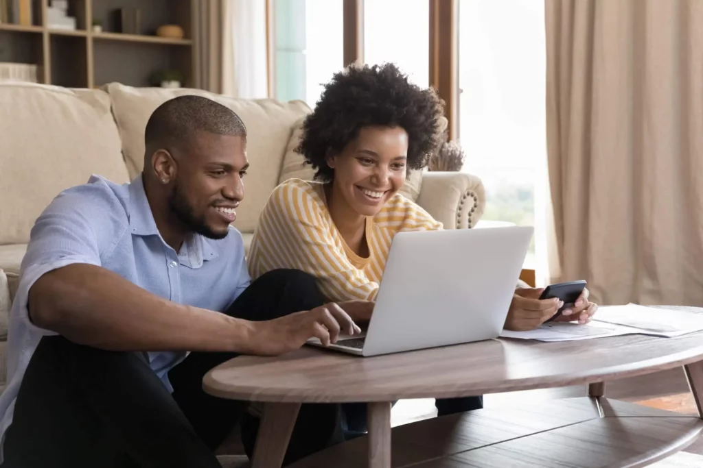 Imagem de um homem e uma mulher sentados no chão de uma sala entre um sofá e uma mesa de centro analisando as informações em uma tela de notebook para ilustrar matéria sobre o contrato de compra e venda com interveniente quitante