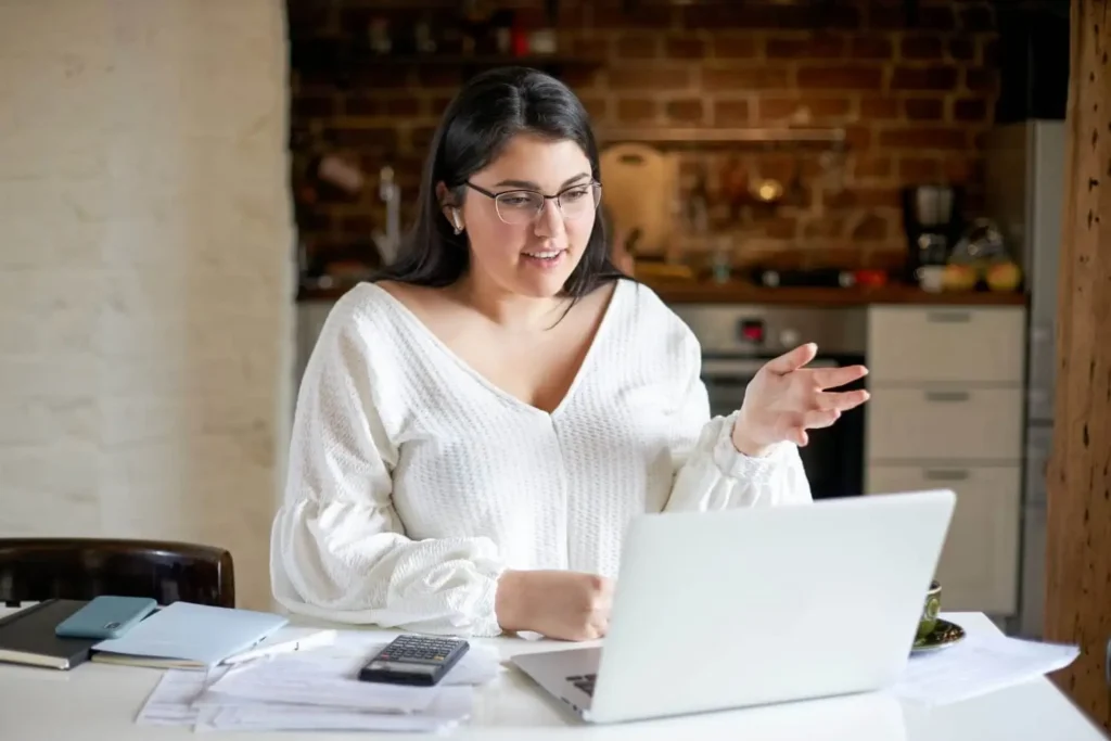 Imagem de uma mulher de cabelos longos e escuros e óculos sentada à mesa, com papéis, uma calculadora e um notebook, gesticulando enquanto faz uma videochamada em uma cozinha aconchegante com tijolos à vista, para ilustrar matéria sobre como escolher um inquilino.