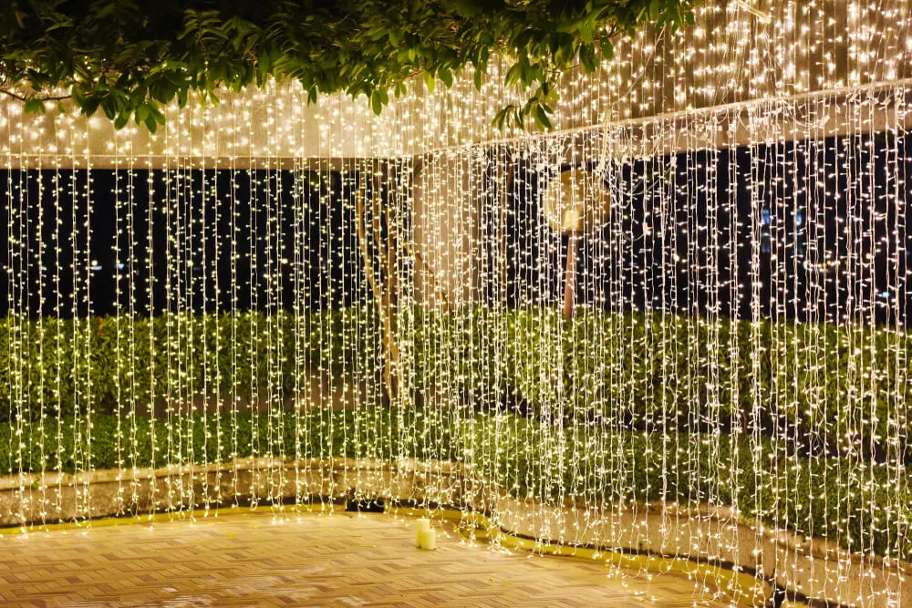 Imagem de uma área externa decorada com muitas luzes de corda penduradas, criando uma cortina de luzes brancas quentes. Arbustos e árvores verdes são visíveis ao fundo, e o chão é pavimentado com ladrilhos.