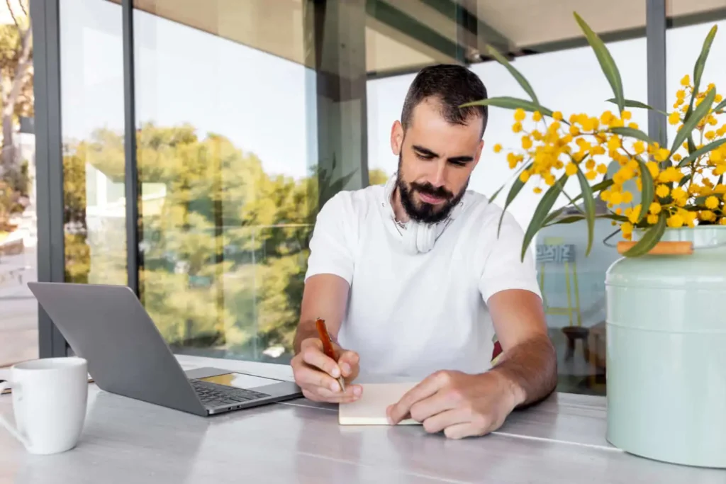Imagem de um homem com fones de ouvido pendurados no pescoço escreve em um caderno em uma mesa onde há um notebook, uma caneca e um vaso com flores amarelas, sentado em uma sala bem iluminada e moderna com janelas amplas.