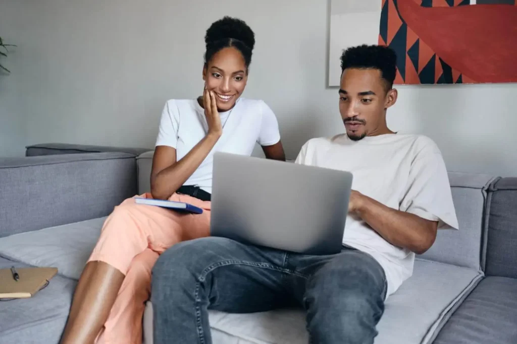 Imagem de uma mulher sorridente e um homem sentados juntos em um sofá cinza, olhando para um notebook. Os dois parecem envolvidos e concentrados. Uma pintura colorida está pendurada na parede atrás deles.