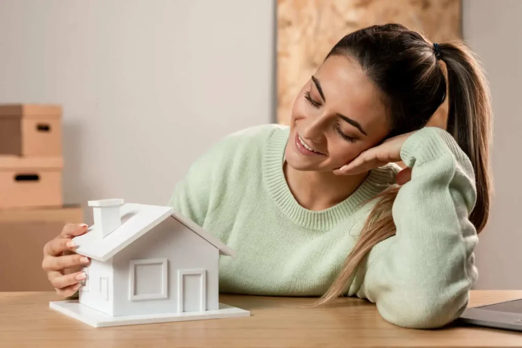 Imagem de uma mulher com o braço encostado em uma mesa enquanto olha para uma casa de madeira em tamanho miniatura que está em cima da mesa para ilustrar matéria sobre como comprar apartamento sem entrada