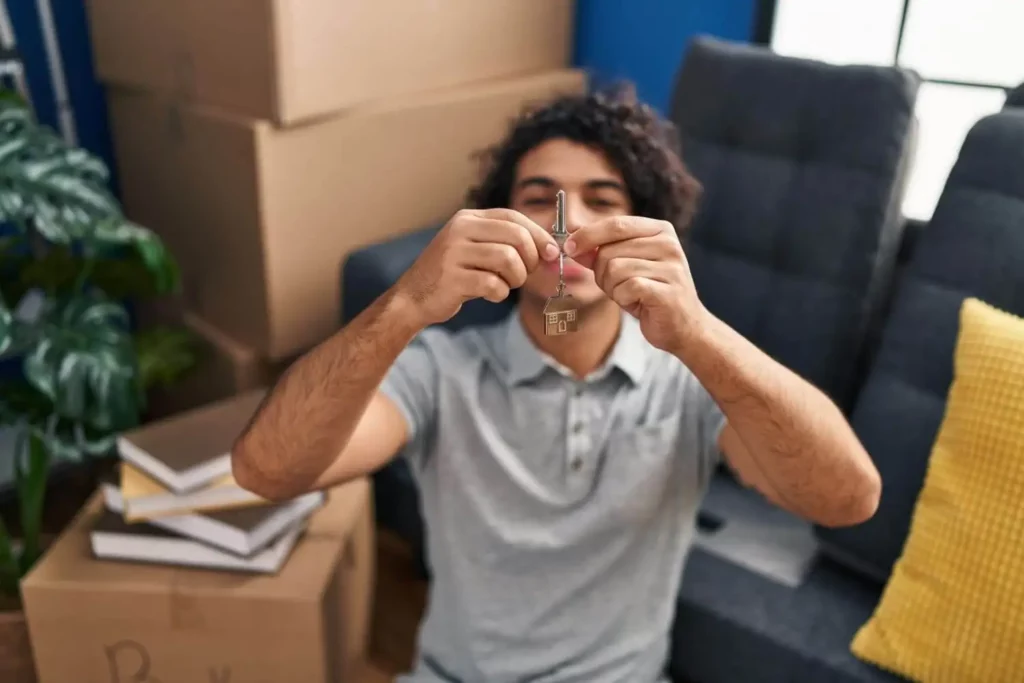 Imagem de um homem segurando a chave de um casa dentro de uma sala de estar decorada com um sofá, plantas, almofada e algumas caixas de papelão para ilustrar matéria sobre como comprar imóvel sem entrada
