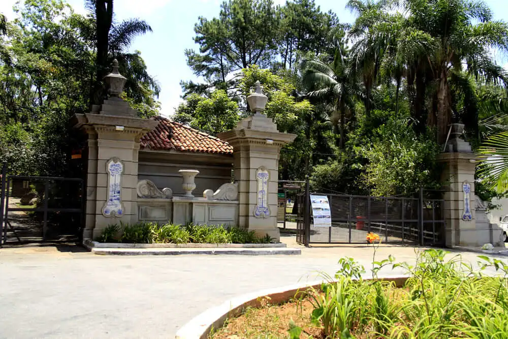 Imagem de um portão de entrada de pedra com pilares decorativos, detalhes em azulejos e uma estrutura de telhado com telhas vermelhas ao fundo, no Horto Florestal, um dos parques em São Paulo. 