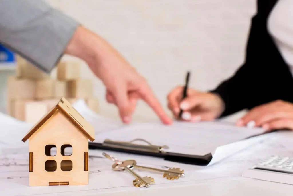 Imagem de uma pessoa apontando para um documento que está em cima de uma mesa ao lado de um modelo de casa em miniatura e um molho de chaves.
