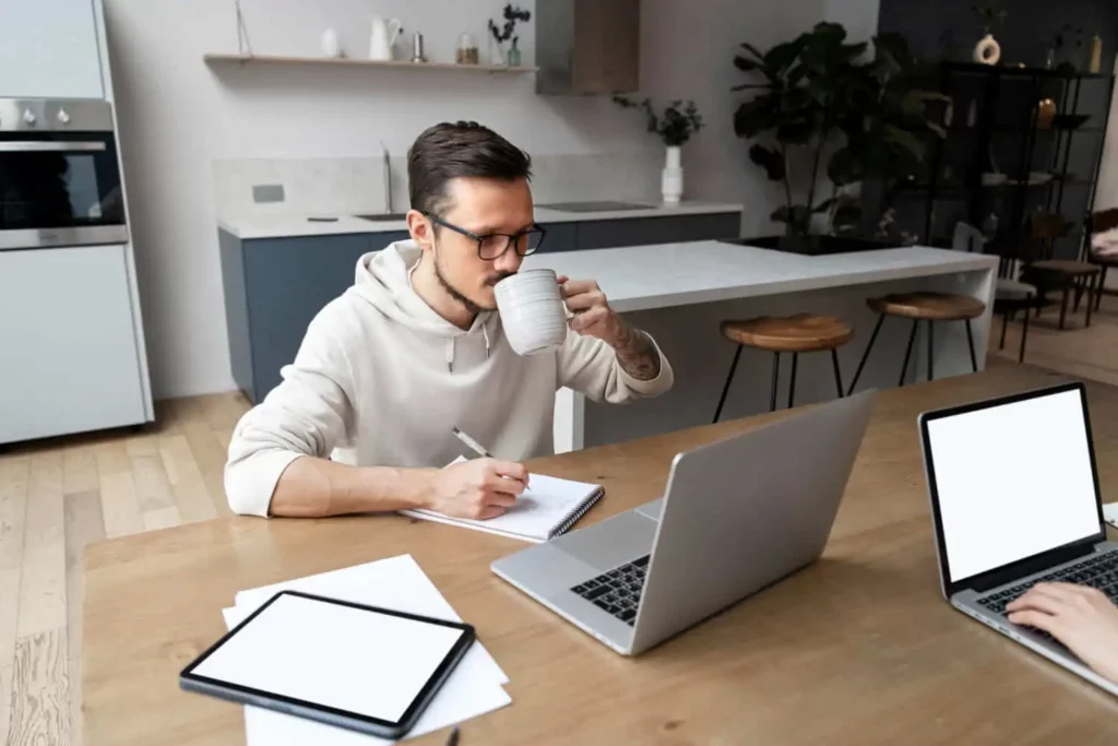 Imagem de um homem sentado em uma mesa com um laptop, um bloco de notas e um tablet, tomando um gole de uma caneca e escrevendo anotações do ITCMD. Outro laptop e papéis estão visíveis sobre a mesa em uma cozinha moderna.