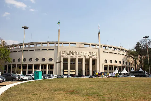 Imagem do Estádio Municipal Paulo Machado de Carvalho, mais conhecido como Estádio do Pacaembu, com vários mastros, carros estacionados e pessoas reunidas perto de barracas