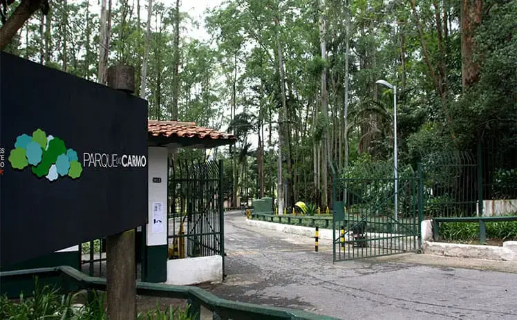 Imagem da fachada de entrada do Parque do Carmo, em SP, mostra o portão de entrada, uma placa com o nome do espaço e árvores ao fundo
