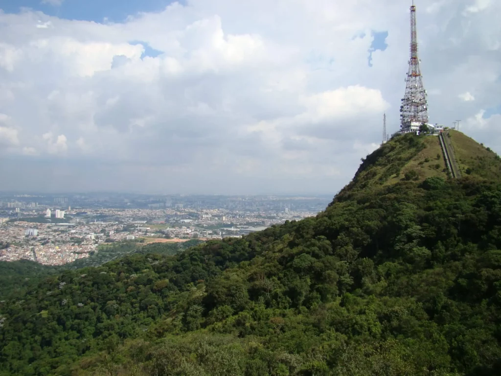 Imagem de uma grande torre de rádio no topo de uma colina verde e arborizada do Pico do Jaraguá, um dos principais parques públicos em São Paulo. 