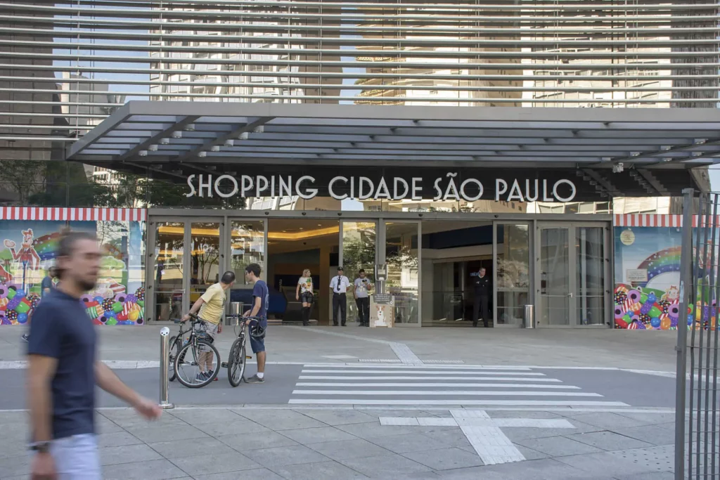 Imagem de pessoas caminhando e andando de bicicleta em frente à entrada do Shopping Cidade São Paulo, um shopping moderno com portas de vidro e decorações coloridas 