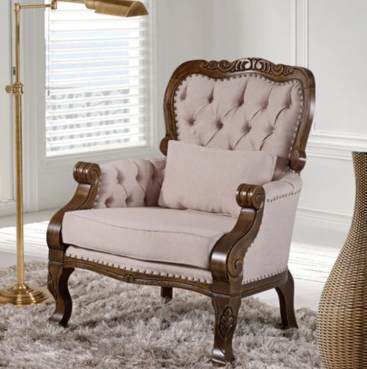 Uma poltrona de estilo vintage com estrutura de madeira ornamentada e estofamento capitonê rosa claro, além de uma almofada combinando, fica sobre um tapete felpudo ao lado de uma luminária de chão dourada em uma sala iluminada e elegante.
