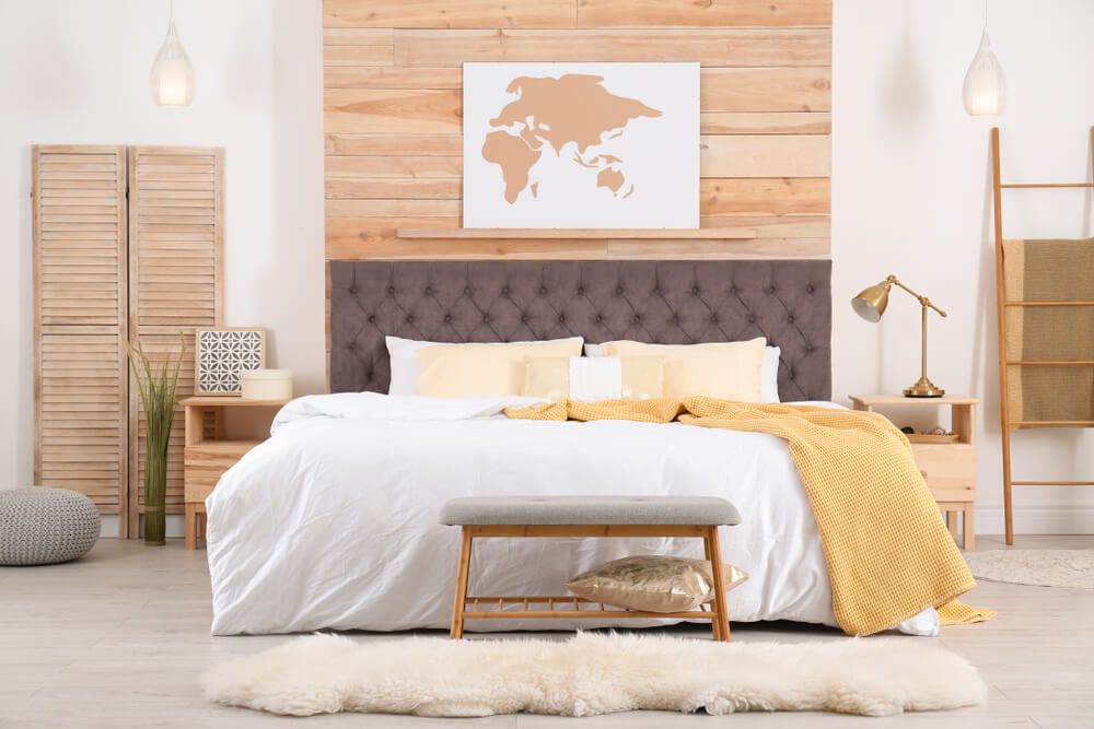 Um quarto moderno com uma cama grande com roupa de cama branca e amarela, cabeceira acolchoada em Capitonê, detalhes em madeira e uma arte de mapa-múndi acima da cama. Elementos de decoração aconchegantes e iluminação suave completam o espaço convidativo.