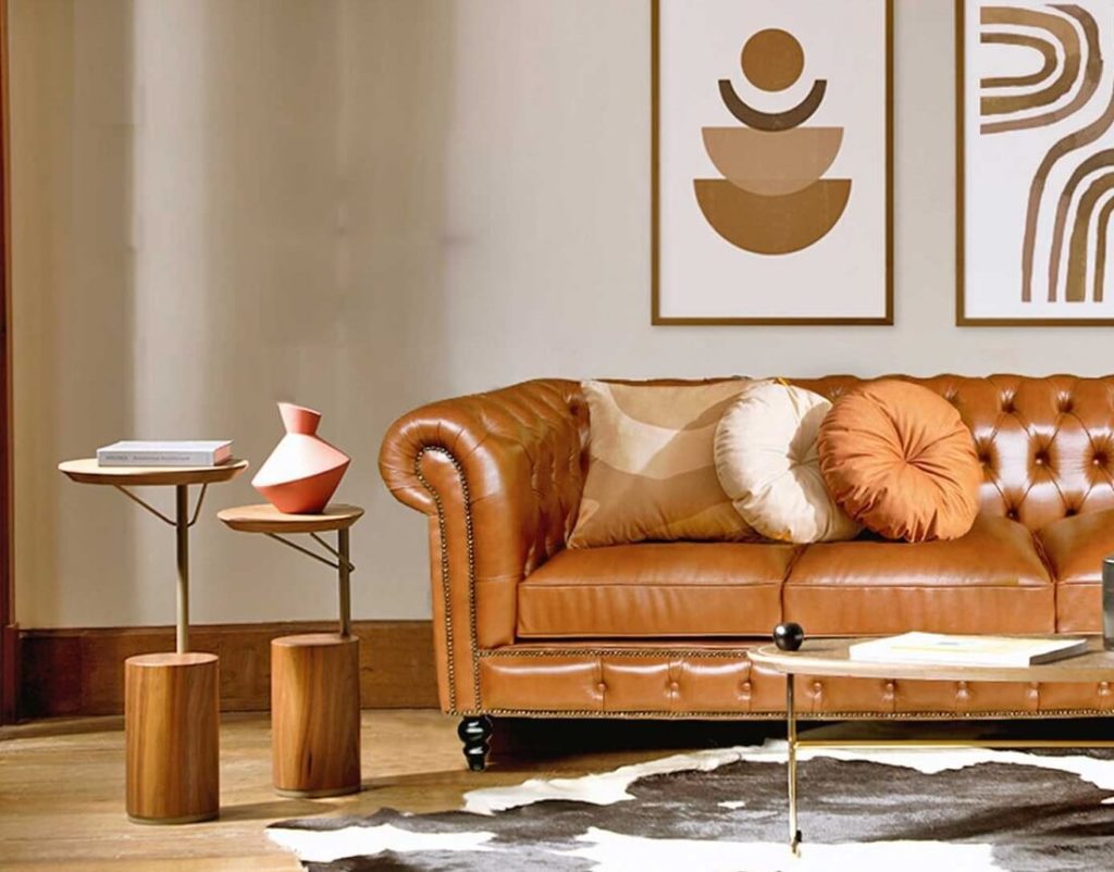 Uma sala de estar elegante com um sofá de couro capitonê marrom, almofadas redondas e quadradas, mesas laterais de madeira modernas, arte abstrata na parede e um tapete de couro preto e branco sobre um piso de madeira.