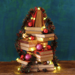 Uma pilha de livros dispostos em formato triangular, como uma árvore de Natal, decorada com enfeites de Natal, enfeites e luzes de Natal — inspiração perfeita para seu próximo quiz de natal — sobre um fundo azul esverdeado em uma superfície de madeira.