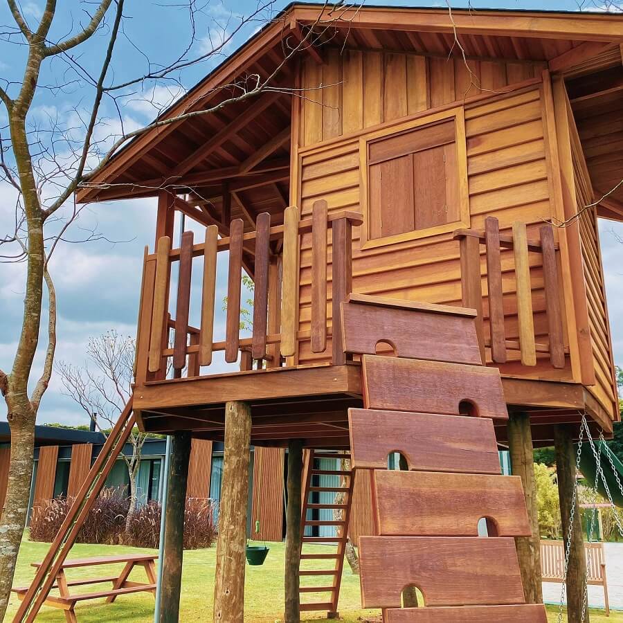 Uma casa de madeira na árvore simples, com uma escada e varanda cercada, fica sobre palafitas em um quintal gramado, com uma árvore, mesa de piquenique e balanço por perto, sob um céu parcialmente nublado.