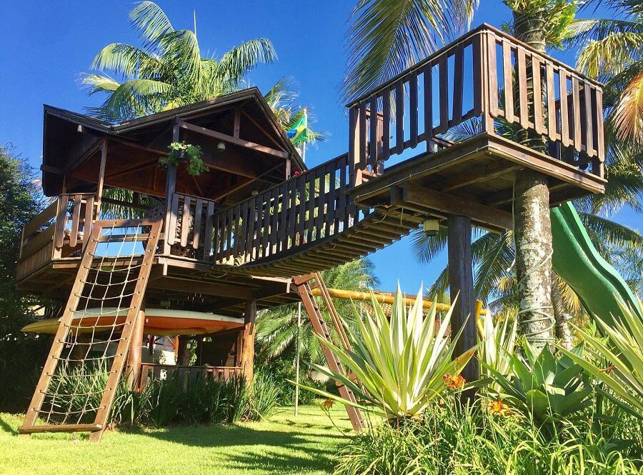 Uma grande casa de madeira na árvore simples, com uma ponte, rede de escalada de corda, escorregador e varanda, fica entre palmeiras e vegetação exuberante sob um céu azul brilhante.