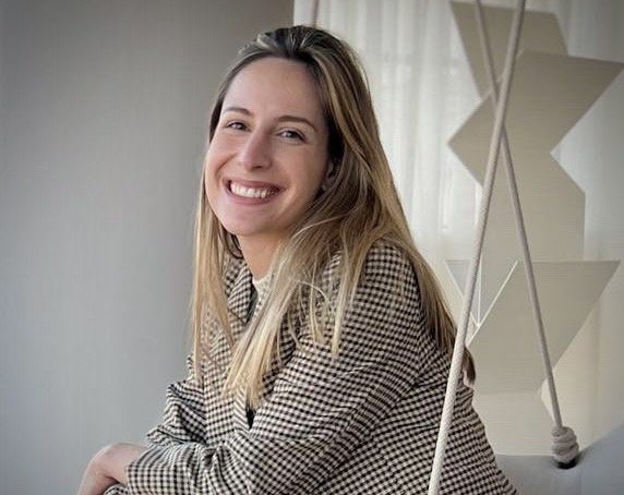 Uma mulher com longos cabelos loiros, vestindo um blazer xadrez, sorri sentada em um balanço dentro de casa, cercada por uma luz suave e decoração geométrica boho ao fundo.