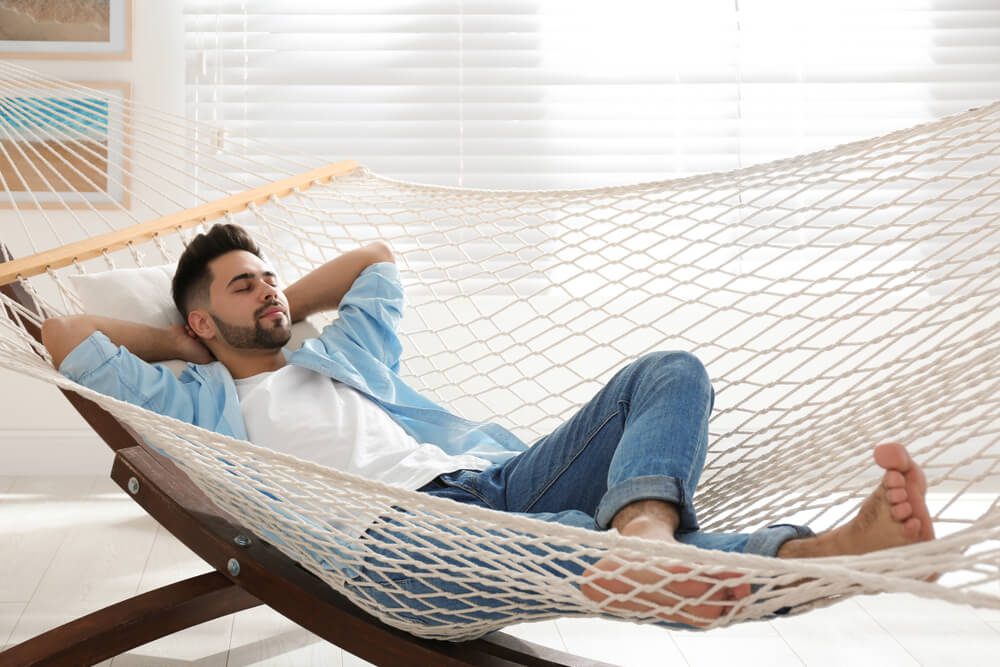 Um homem barbudo relaxa em uma sala de descanso, deitado em uma rede branca, com as mãos atrás da cabeça e os olhos fechados. Ele veste uma camisa azul-clara e calça jeans, enquanto a luz do sol penetra pelas persianas ao fundo.