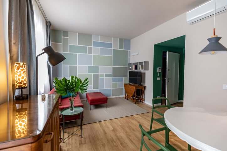 Uma sala de estar moderna com uma parede geométrica, sofá vermelho, pufe, cadeiras verdes, uma pequena mesa de jantar redonda, plantas de interior, pisos de madeira e grandes janelas com cortinas transparentes.